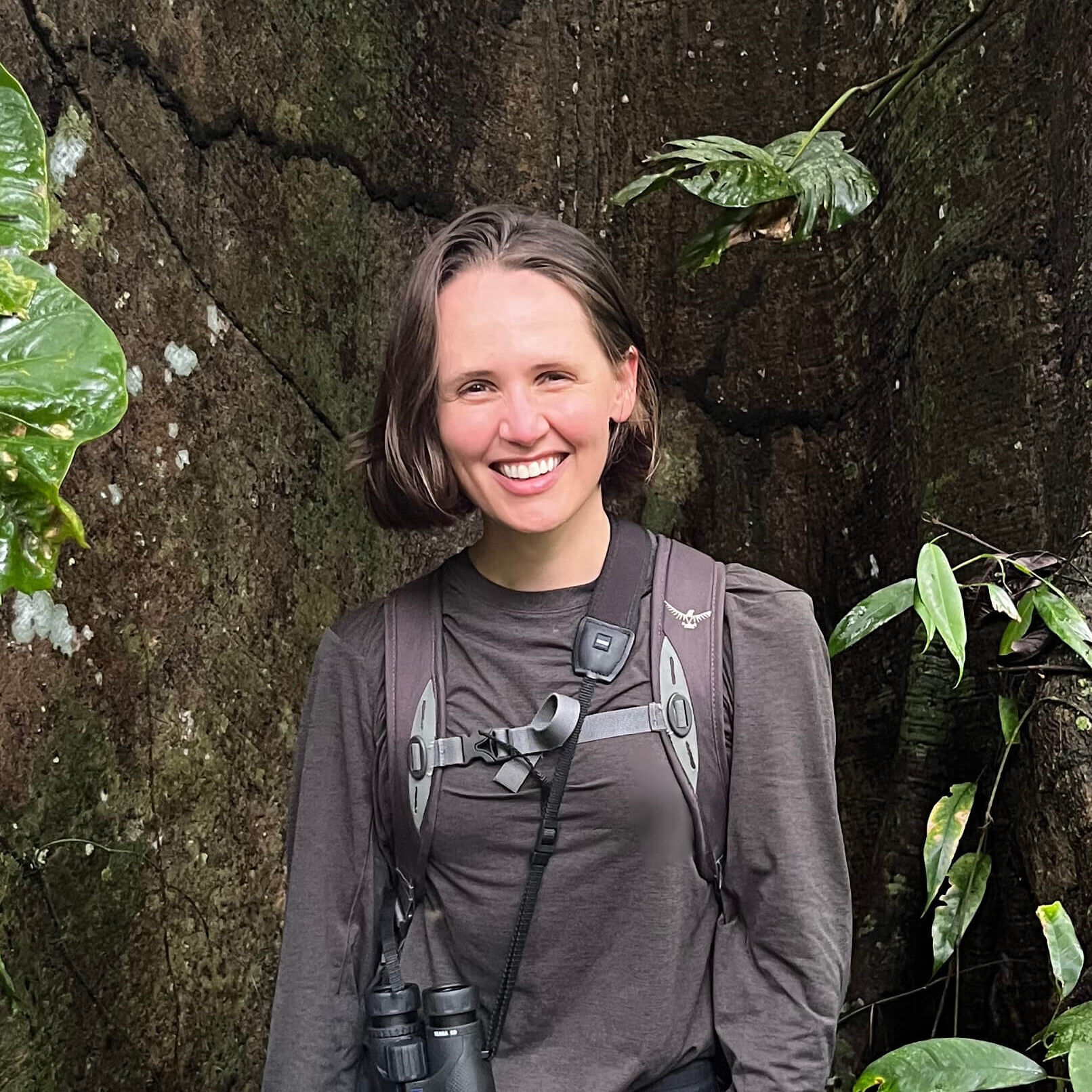 Professional headshot of Della Fetzer, smiling outdoors in front of a stone wall with green foliage. She is wearing a gray long-sleeve shirt and a backpack.