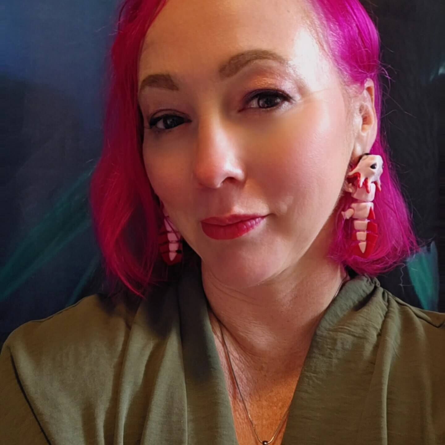 Professional headshot of Erin Horton, a woman with bright pink hair and long earrings, smiling and wearing a forest green shirt.
