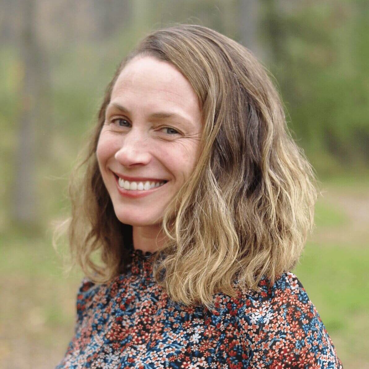 Portrait of Jill Dohner smiling, wearing a colorful patterned blouse against a soft green natural outdoor background.