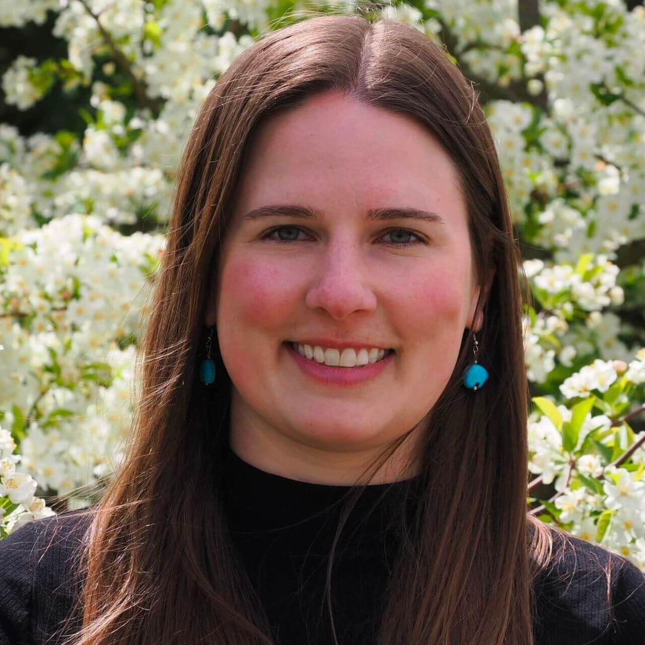 Close-up headshot of Kelly Krueger smiling with brown hair and turquoise earrings against a background of white flowering blossoms.
