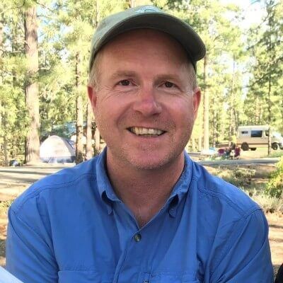 Professional headshot of Mike Smalligan, a man with a grey baseball cap and blue shirt, smiling in an outdoor wooded setting with trees and a tent in the background.