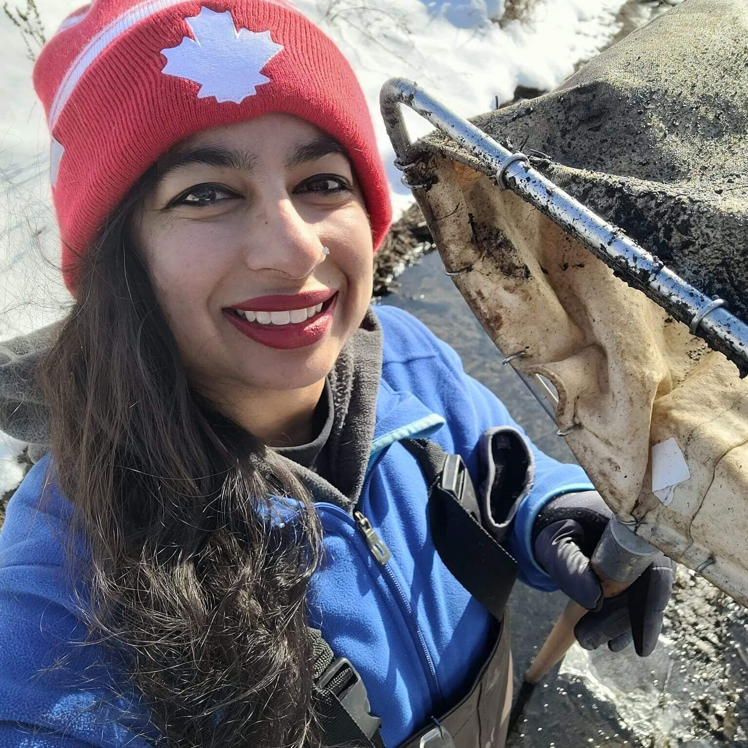 Dr. Shikha Singh, JLW CISMA Coordinator, smiling in the field wearing a red Canada winter hat and holding a professional aquatic sampling net.